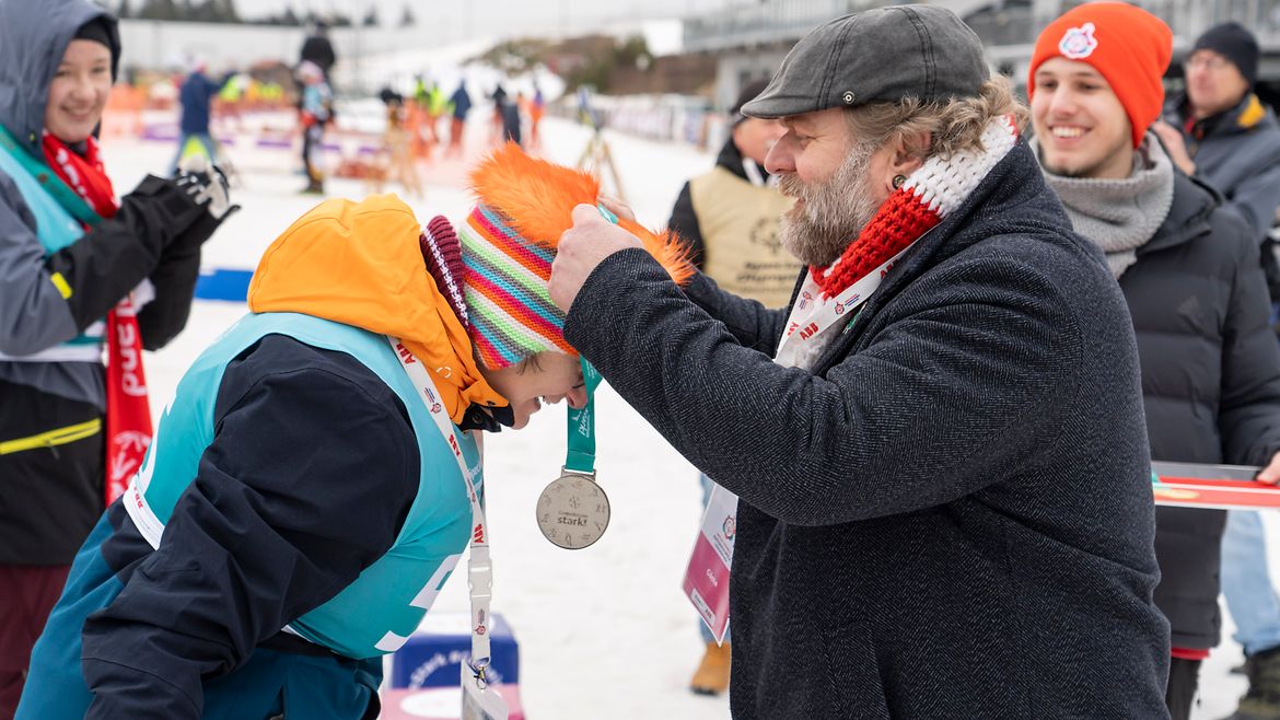 Ein Mann mit Mütze und Winterjacke hängt einer Athletin mit Mütze und Winterjacke eine Silbermedaille um. Im Hintergrund weitere Personen.