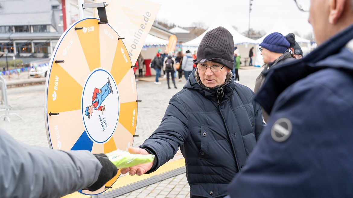 3 Personen vor dem BGW-Glücksrad mit der Figur Nils drauf. Ein Arm reicht der Person in der Mitte einen BGW-Gewinn.