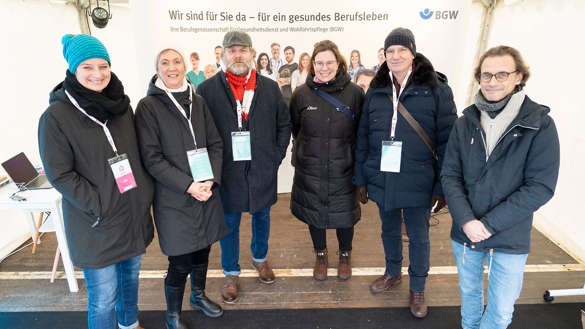 3 Frauen und 3 Männer stehen nebeneinander in Winterjacken vor dem BGW-Messestand. Im Hintergrund ein großes Poster mit verschiedenen Personen und dem Slogan "Wir sind für Sie da - für ein gesundes Berufsleben" und BGW-Logo.