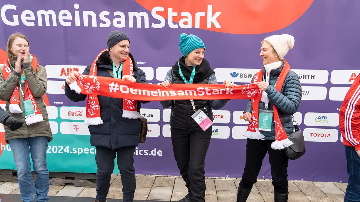 4 Frauen nebeneinander vor einer Wand mit diversen Firmen-Logos und dem Slogan "GemeinsamStark". 3 der Frauen halten zusammen einen Schal mit der Aufschrift "#GemeinsamStark".